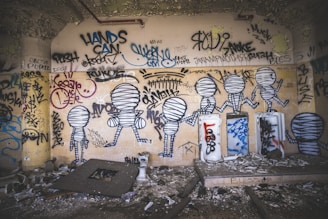 A wall covered with various graffiti and street art, including abstract figures resembling mummified humans. The drawings are in black and white, surrounded by tags and colorful scribbles. The room is dimly lit, with a gritty and decayed environment, featuring debris and peeling paint.