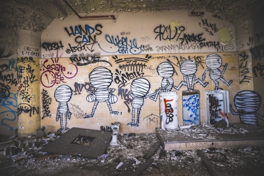 A wall covered with various graffiti and street art, including abstract figures resembling mummified humans. The drawings are in black and white, surrounded by tags and colorful scribbles. The room is dimly lit, with a gritty and decayed environment, featuring debris and peeling paint.