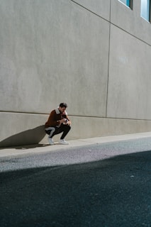A man crouches low, holding a camera in his hands, against the backdrop of a large concrete wall. He is dressed in a brown jacket, black track pants with white stripes, and white sneakers. The scene is set on a city street with the wall casting a long shadow on the ground. The lighting suggests a bright, sunny day.