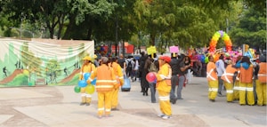 A vibrant outdoor event with people dressed in orange and yellow uniforms holding colorful balloons. A festive backdrop with a green illustrated mural and an arch made of multicolored balloons. A diverse crowd gathers, some holding signs and various objects. Trees and greenery surround the area, giving a lively and community-oriented atmosphere.