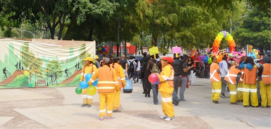 A vibrant outdoor event with people dressed in orange and yellow uniforms holding colorful balloons. A festive backdrop with a green illustrated mural and an arch made of multicolored balloons. A diverse crowd gathers, some holding signs and various objects. Trees and greenery surround the area, giving a lively and community-oriented atmosphere.