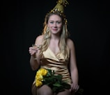 A woman wearing a gold party hat and a golden dress holds a wine glass and a bouquet of yellow roses against a dark background. Her expression is cheerful, and her long hair cascades over her shoulders.