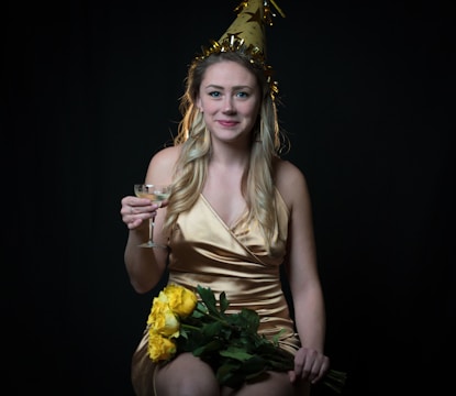 A woman wearing a gold party hat and a golden dress holds a wine glass and a bouquet of yellow roses against a dark background. Her expression is cheerful, and her long hair cascades over her shoulders.