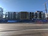 A multi-story building under construction with scaffolding in place. The structure features a modern design with a combination of large glass windows and darker paneling. The construction site is enclosed by a blue barrier, and there is a clear sky in the background with a few trees visible on the left side.