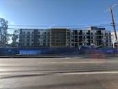 A multi-story building under construction with scaffolding in place. The structure features a modern design with a combination of large glass windows and darker paneling. The construction site is enclosed by a blue barrier, and there is a clear sky in the background with a few trees visible on the left side.