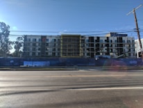 A multi-story building under construction with scaffolding in place. The structure features a modern design with a combination of large glass windows and darker paneling. The construction site is enclosed by a blue barrier, and there is a clear sky in the background with a few trees visible on the left side.