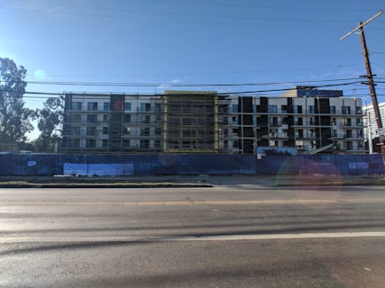 A multi-story building under construction with scaffolding in place. The structure features a modern design with a combination of large glass windows and darker paneling. The construction site is enclosed by a blue barrier, and there is a clear sky in the background with a few trees visible on the left side.
