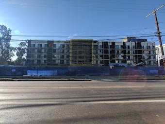 A multi-story building under construction with scaffolding in place. The structure features a modern design with a combination of large glass windows and darker paneling. The construction site is enclosed by a blue barrier, and there is a clear sky in the background with a few trees visible on the left side.