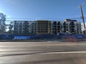 A multi-story building under construction with scaffolding in place. The structure features a modern design with a combination of large glass windows and darker paneling. The construction site is enclosed by a blue barrier, and there is a clear sky in the background with a few trees visible on the left side.