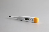 A digital thermometer lying on a light gray surface. It features a white body with an orange cap at one end, and a small LCD display screen near the tip.