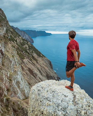 man in blue shirt and black shorts standing on rock formation near body of water during