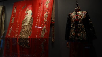 A richly embroidered red textile features intricate designs and figures, including a standing figure in traditional attire. To the right, a mannequin displays a dark, intricately patterned jacket with floral motifs and a similar traditional style.