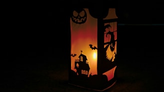 A lantern with intricate Halloween-themed cutouts projects an eerie glow. The design includes a haunted house, bats, and a sinister face, surrounded by a warm, orange-yellow light that creates a spooky atmosphere.