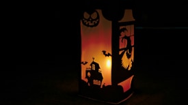 A lantern with intricate Halloween-themed cutouts projects an eerie glow. The design includes a haunted house, bats, and a sinister face, surrounded by a warm, orange-yellow light that creates a spooky atmosphere.