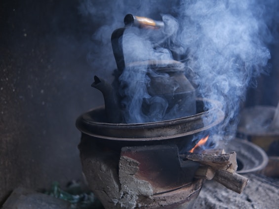 A black kettle is placed on a traditional clay stove with a fire burning beneath it. Smoke billows around the kettle, creating a rustic and cozy atmosphere. The stove appears weathered and well-used, with visible cracks and soot marks.
