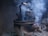 A black kettle is placed on a traditional clay stove with a fire burning beneath it. Smoke billows around the kettle, creating a rustic and cozy atmosphere. The stove appears weathered and well-used, with visible cracks and soot marks.