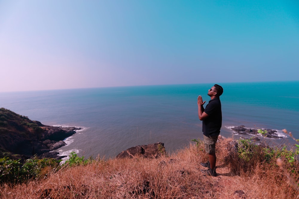 man in black t-shirt standing on brown rock formation near blue sea during daytime