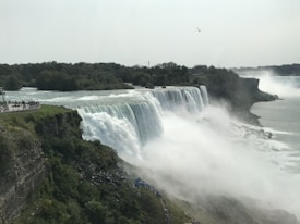 A powerful waterfall with multiple cascades flows over a rocky cliff surrounded by lush greenery. Mist rises from the water, filling the air. On the left, a group of tourists stand on a viewing platform, observing the natural spectacle. A single bird is visible in the sky.