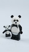 Two panda-themed figurines made of plastic are positioned side by side on a plain white surface. The larger figure has a black and white color scheme with a distinct panda bear face and body. It holds the smaller figure's hand, which also resembles a panda with similar features. The figures have exaggerated, toy-like proportions, with the larger figure featuring more noticeable articulation and blocky limbs.
