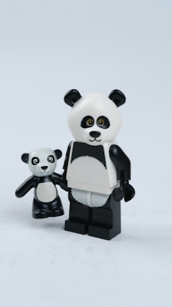 Two panda-themed figurines made of plastic are positioned side by side on a plain white surface. The larger figure has a black and white color scheme with a distinct panda bear face and body. It holds the smaller figure's hand, which also resembles a panda with similar features. The figures have exaggerated, toy-like proportions, with the larger figure featuring more noticeable articulation and blocky limbs.