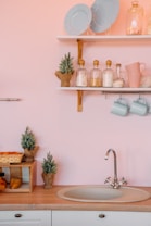 A cozy kitchen setting with a sink, two shelves, and decorative elements. On the top shelf, there are two light blue plates and glass jars, one with a small Christmas tree, and a ceramic mug. The lower shelf holds three glass jars containing various items, a pink mug, and two light blue hanging mugs. Below, there is a sink with a clean faucet on a wooden countertop, surrounded by small potted pine trees and a bread basket.