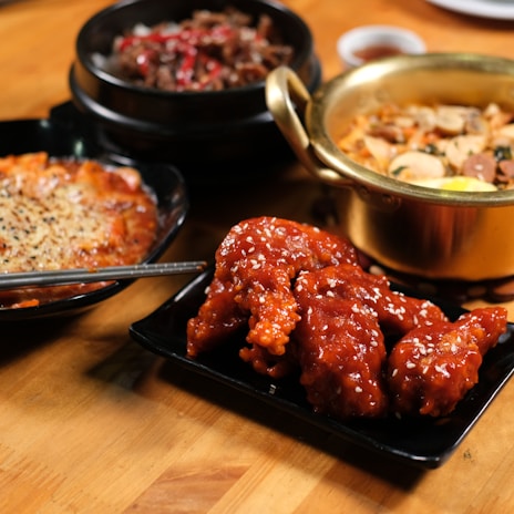 A variety of Korean dishes are presented on a wooden table. The foreground features a plate of glazed, sesame-seeded fried chicken wings. In the background, there are a bowl of spicy beef stew with vegetables, a golden pot containing a soup or stew with sliced ingredients, and a dish with melted cheese on top.