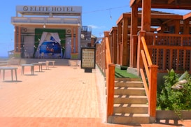 An outdoor area of a hotel with a large sign reading 'Elite Hotel' at the top. The open space is covered with light-colored tiles, leading to a stage area with a backdrop image of open seas. There are several small round tables scattered across the open area. To the right, there's a wooden structure with steps, decorated with green turf and plants.