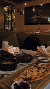 A warmly lit dining setup featuring a wooden table with various dishes including a flatbread, cooked vegetables in small pots, and side condiments. Soft candlelight enhances the cozy atmosphere, and plush seating lines one side of the table. A large window and a painting of a ship adorn the walls, while glasses and a single flower on a table add to the elegant decor.