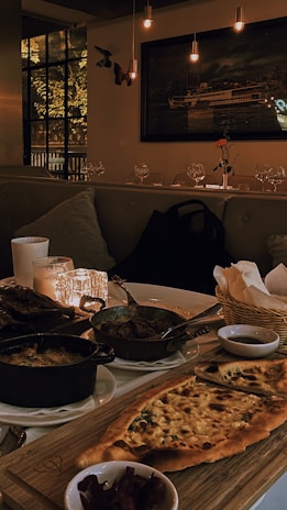 A warmly lit dining setup featuring a wooden table with various dishes including a flatbread, cooked vegetables in small pots, and side condiments. Soft candlelight enhances the cozy atmosphere, and plush seating lines one side of the table. A large window and a painting of a ship adorn the walls, while glasses and a single flower on a table add to the elegant decor.