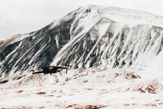 A drone is flying in front of a large, snowy mountain. The rugged mountain features a mix of snow and exposed rock, creating a striking contrast. The landscape is vast and appears to be in a remote area.