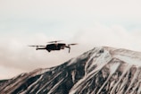 A drone hovers in the foreground against a backdrop of a snow-covered mountain. The mountain exhibits shades of grey and white with bold angular ridges, under a cloudy sky.