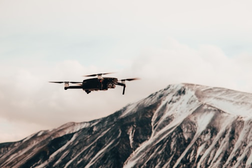 A drone hovers in the foreground against a backdrop of a snow-covered mountain. The mountain exhibits shades of grey and white with bold angular ridges, under a cloudy sky.