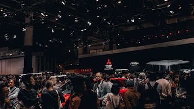 A crowded exhibition hall featuring people gathered around several cars on display. The background shows large Mitsubishi Motors logos and 'K-Wagon Concept' signage. Overhead lights illuminate the scene, enhancing the vibrant and energetic ambiance of the event.