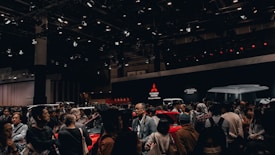 A crowded exhibition hall featuring people gathered around several cars on display. The background shows large Mitsubishi Motors logos and 'K-Wagon Concept' signage. Overhead lights illuminate the scene, enhancing the vibrant and energetic ambiance of the event.