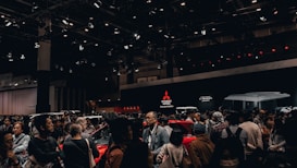 A crowded exhibition hall featuring people gathered around several cars on display. The background shows large Mitsubishi Motors logos and 'K-Wagon Concept' signage. Overhead lights illuminate the scene, enhancing the vibrant and energetic ambiance of the event.