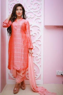 A woman stands elegantly in a pink and orange traditional outfit, featuring intricate patterns. She has long hair, adorned with gold earrings, and is gracefully holding a matching dupatta. The background is a soft pink with ornate, white patterns.