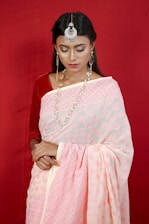 A woman with long dark hair is wearing a traditional pink and white sari against a red background. She accessorizes with large silver earrings, a striking headpiece, and a layered pearl necklace. Her eyes are closed, and she carries a serene expression.