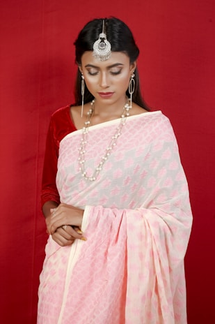 A woman with long dark hair is wearing a traditional pink and white sari against a red background. She accessorizes with large silver earrings, a striking headpiece, and a layered pearl necklace. Her eyes are closed, and she carries a serene expression.
