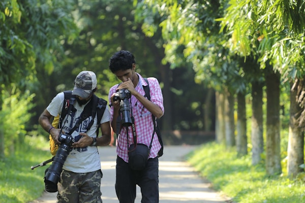Two people are walking along a sunny pathway lined with trees, each holding a camera. They appear to be focused on their photography activities, possibly reviewing photos they’ve taken.