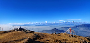 Vast mountainous landscape featuring snow-capped peaks in the distance under a clear blue sky. A small building, possibly a stupa, is situated on a grassy hill with prayer flags draped around it. Another set of colorful prayer flags is arranged in a conical shape on the right side of the image.