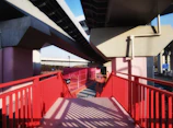 A modern walkway under elevated highway structures, featuring bold red railings and pink support columns. Shadows create patterns on the walkway, adding depth and interest. The sky is clear, and the architectural forms appear geometric and industrial.