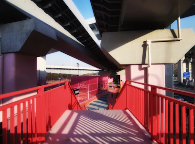 A modern walkway under elevated highway structures, featuring bold red railings and pink support columns. Shadows create patterns on the walkway, adding depth and interest. The sky is clear, and the architectural forms appear geometric and industrial.