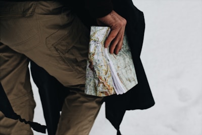 A person wearing beige pants holds a map in their left hand against a snowy background. The map is detailed, showing various geographical features.