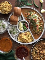 A flavorful Indian meal served on a segmented metal plate includes spicy curries, freshly chopped onions, tangy lemon wedges, and colorful green and red chilies. Accompanying the meal are soft bread rolls and a bowl of namkeen snacks. Green bangles, lemon slices, and whole green chilies decorate the wooden table.