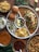 A flavorful Indian meal served on a segmented metal plate includes spicy curries, freshly chopped onions, tangy lemon wedges, and colorful green and red chilies. Accompanying the meal are soft bread rolls and a bowl of namkeen snacks. Green bangles, lemon slices, and whole green chilies decorate the wooden table.