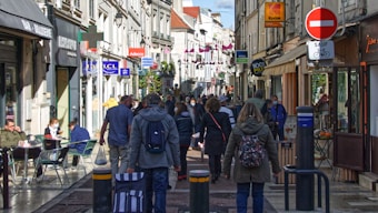 A bustling European street lined with shops and people walking in both directions. Some people are sitting at outdoor café tables on the left side. Various shop signs, including pharmacy and clothing store signs, are visible above the street. Bright sunlight illuminates the scene, and there are barriers indicating a pedestrian-only zone.