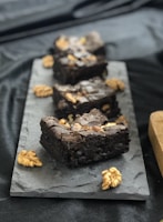 Four pieces of rich, dark chocolate brownies topped with walnuts are arranged on a dark slate serving platter. Whole walnut pieces are scattered around the brownies, enhancing the presentation. The background fabric is a smooth black material, adding a luxurious feel to the setup.