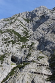 A rugged mountain landscape with steep, rocky slopes features a series of wooden pathways winding up the incline, leading to a large cave entrance. Sparse greenery clings to the rocks, and the sky is clear and blue.