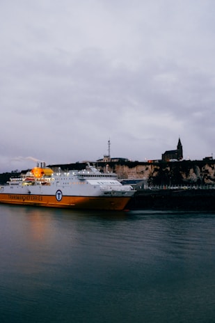 A large ferry with a bright yellow and white exterior is docked at a harbor. The ferry bears the name Transmanche Ferries, and there is a rocky shoreline with buildings atop the cliffs. The sky is filled with gray clouds, and the water reflects the colors of the ferry.