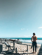 A person with a wetsuit tied around their waist stands on a sandy beach, looking towards the ocean. A surfboard is placed on a wooden bench next to them. The sky is clear and blue, creating a peaceful coastal scene.